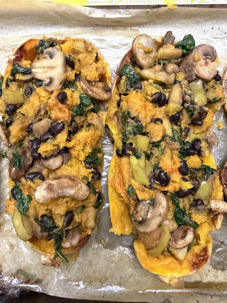 Stuffed butternut squash halves filled with a colorful vegan mixture of mushrooms, black beans, apples, spinach, and roasted squash, shown after baking in the oven.