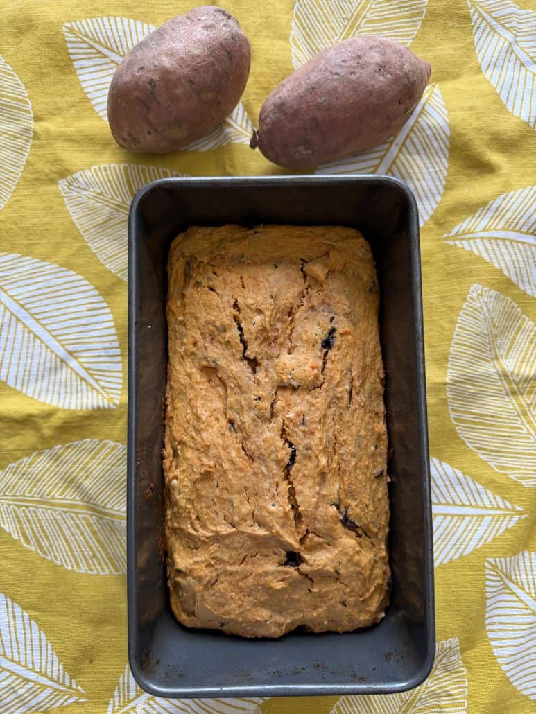 Freshly baked sweet potato bread in a loaf pan, shown with raw sweet potatoes in the background.