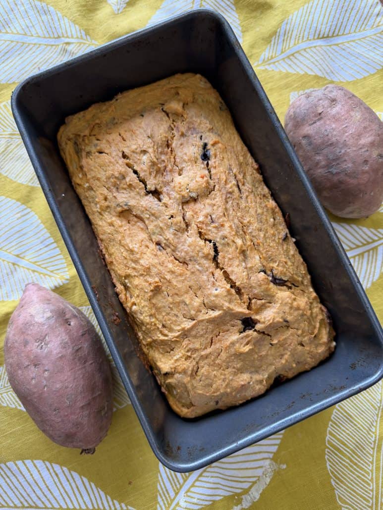 Freshly baked sweet potato bread in a loaf pan, surrounded by whole raw sweet potatoes.