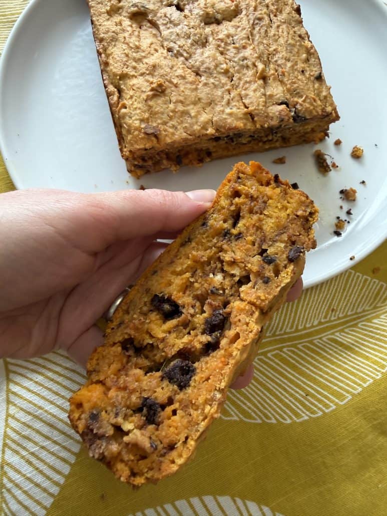 Hand holding a slice of sweet potato chocolate chip bread, showing its soft, moist texture with the full loaf in the background.