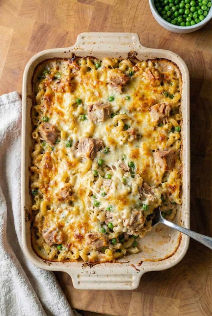 Cheesy tuna casserole being scooped from a baking dish, showing melted cheese, tender pasta, peas, and chunks of tuna.