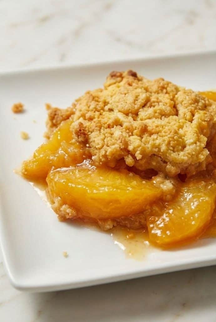 A serving of 3 Ingredient Cake Mix Peach Cobbler on a white plate, showing juicy canned peaches and a golden crumb topping.