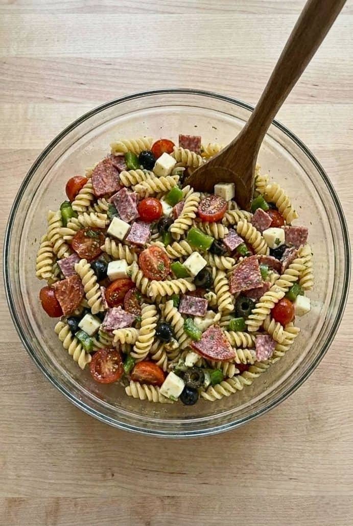 Large glass bowl of freshly mixed Antipasto Pasta Salad with rotini, meats, cheese, olives, and veggies. Ready to chill for a flavorful make-ahead side dish.