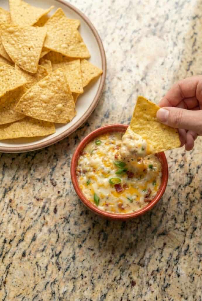 Hand dipping a tortilla chip into a bowl of warm Baked Buffalo Chicken Dip, served with a side of crispy chips.