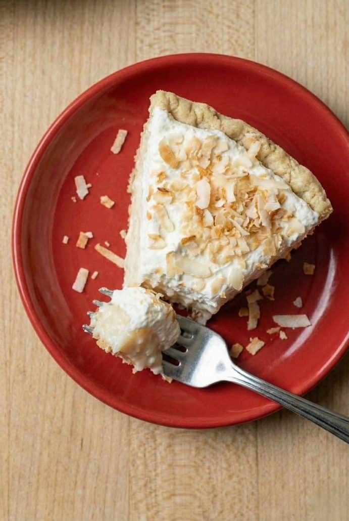 Fork taking a bite from a slice of coconut cream pie on a red plate, showing the creamy filling and toasted coconut topping.