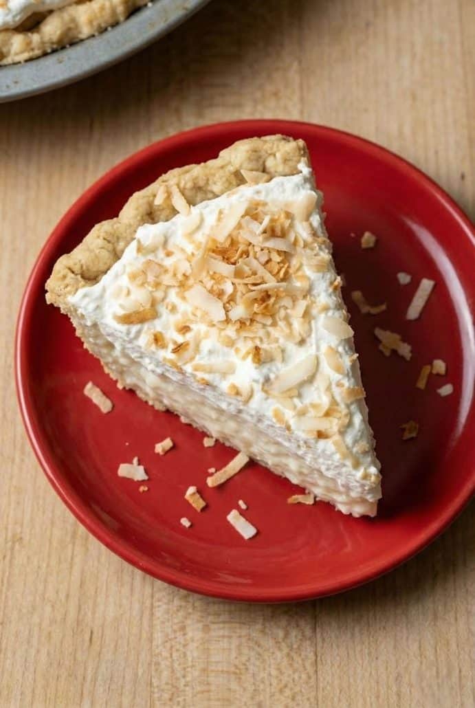 Single slice of coconut cream pie on a red plate, topped with whipped cream and toasted coconut flakes, highlighting the creamy texture.