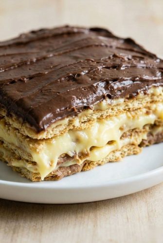 Slice of no-bake éclair cake with layers of graham crackers, vanilla pudding, and chocolate frosting on a white plate.