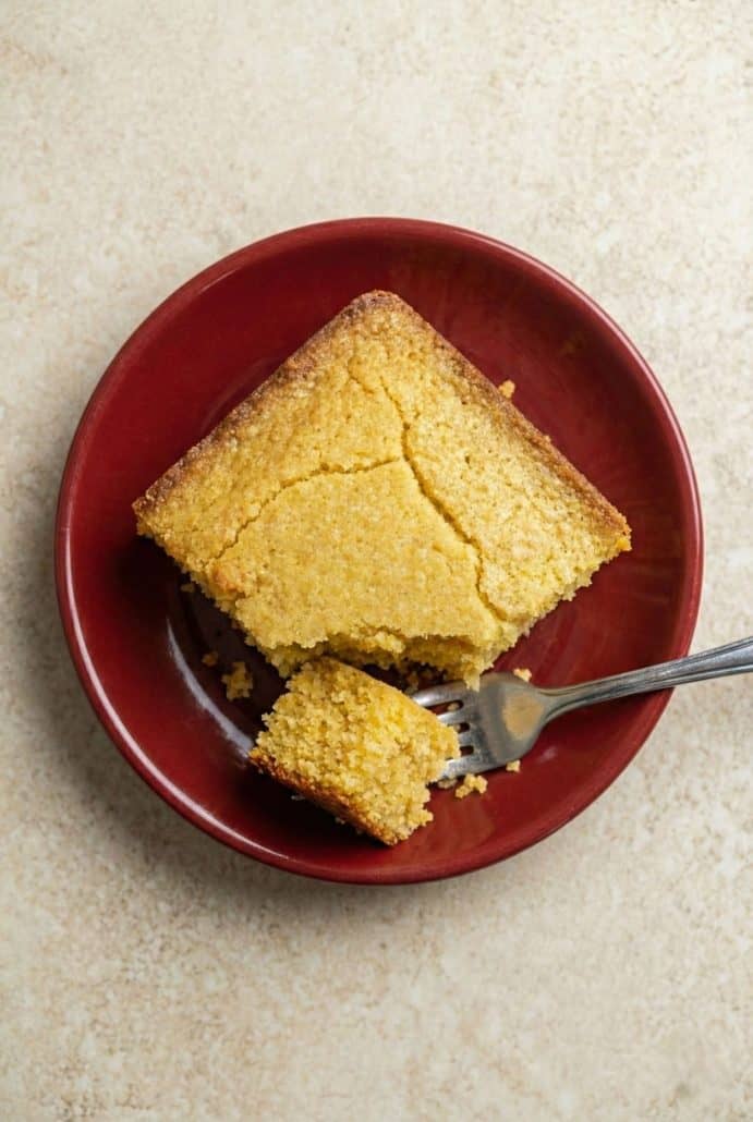 A slice of healthy cornbread on a red plate with a forkful taken out, showing the fluffy texture.