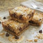 Three gooey Seven Layer Bars stacked on a plate, topped with toasted coconut and chocolate chips.