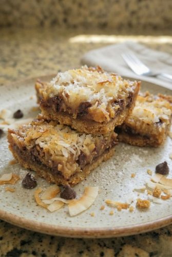 Three gooey Seven Layer Bars stacked on a plate, topped with toasted coconut and chocolate chips.