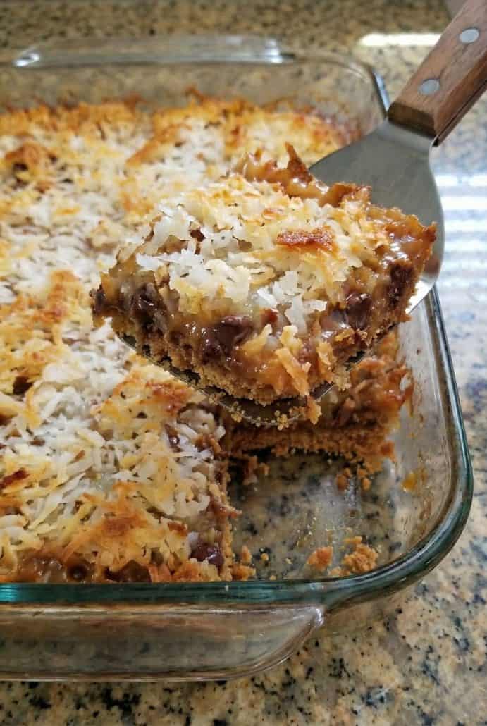 A spatula lifting gooey, golden Seven Layer Bars from a baking dish. Showing the rich, chocolatey layers and toasted coconut topping.