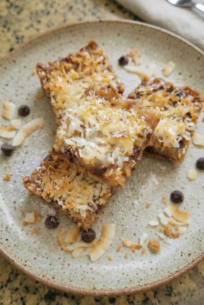 Three Seven Layer Bars on a ceramic plate, topped with toasted coconut and surrounded by chocolate chips.