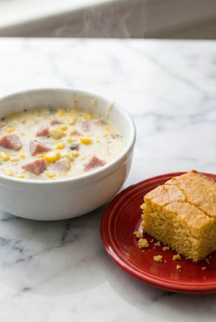 Bowl of slow cooker corn chowder served with a slice of cornbread on a red plate.