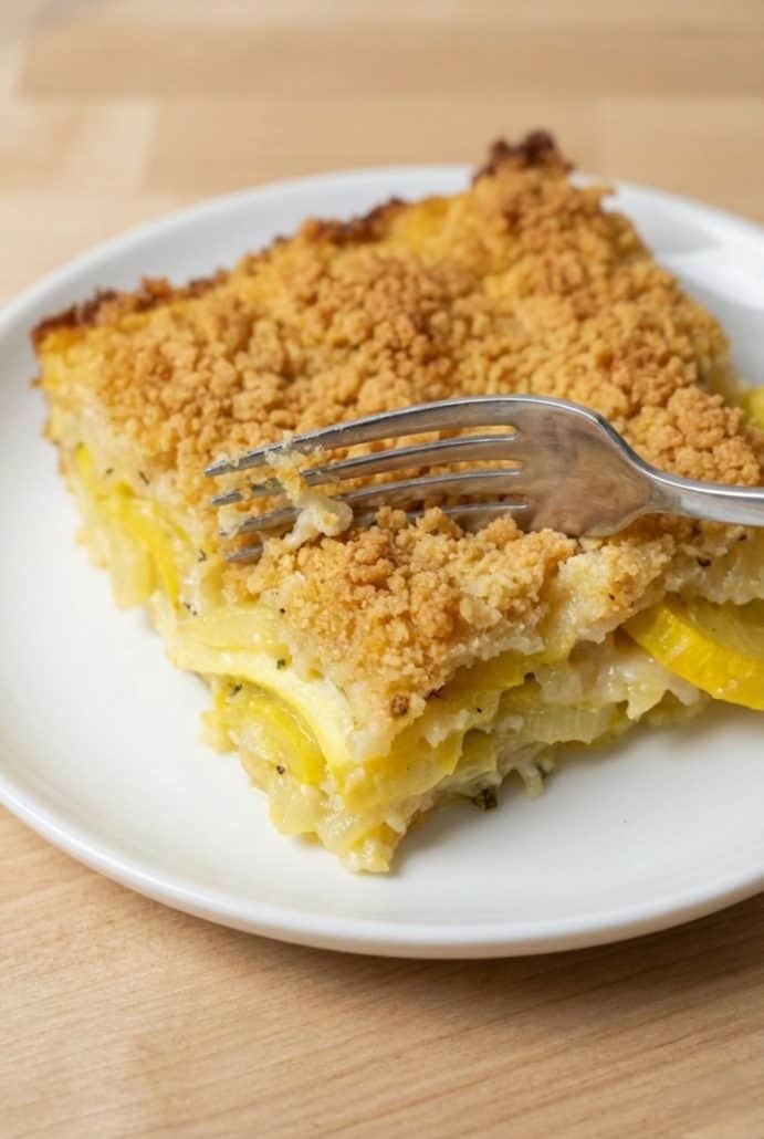 Fork cutting into a warm slice of squash casserole on a white plate, showing the creamy interior and crispy cracker topping.