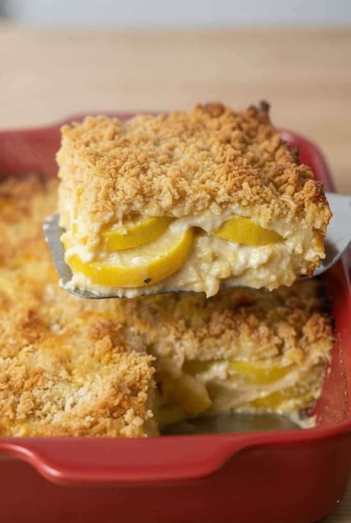 A cheesy slice of baked squash casserole being lifted from the dish, revealing tender layers of yellow squash and a crisp, golden topping.