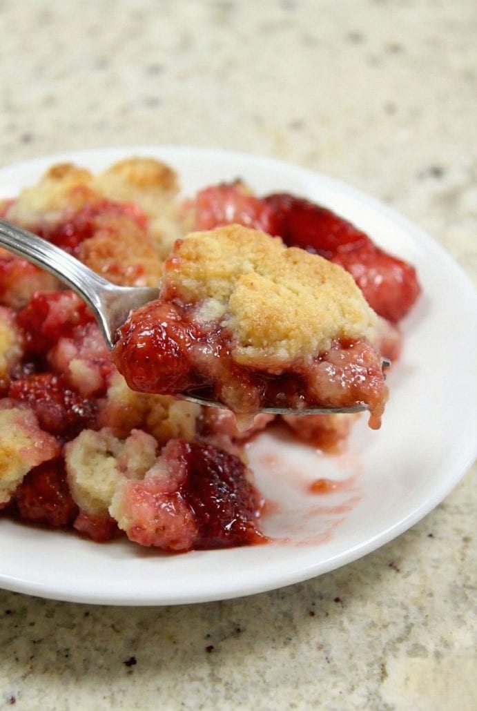 Spoonful of warm strawberry cobbler lifted from a plate, showing gooey fruit filling and golden topping.