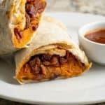 Sweet potato burrito cut in half showing the cheesy bean and sweet potato filling, served on a white plate with dipping sauce.