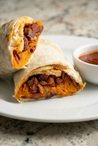 Sweet Potato Burritos - Easy Vegetarian Recipe Sweet potato burrito cut in half showing the cheesy bean and sweet potato filling, served on a white plate with dipping sauce.