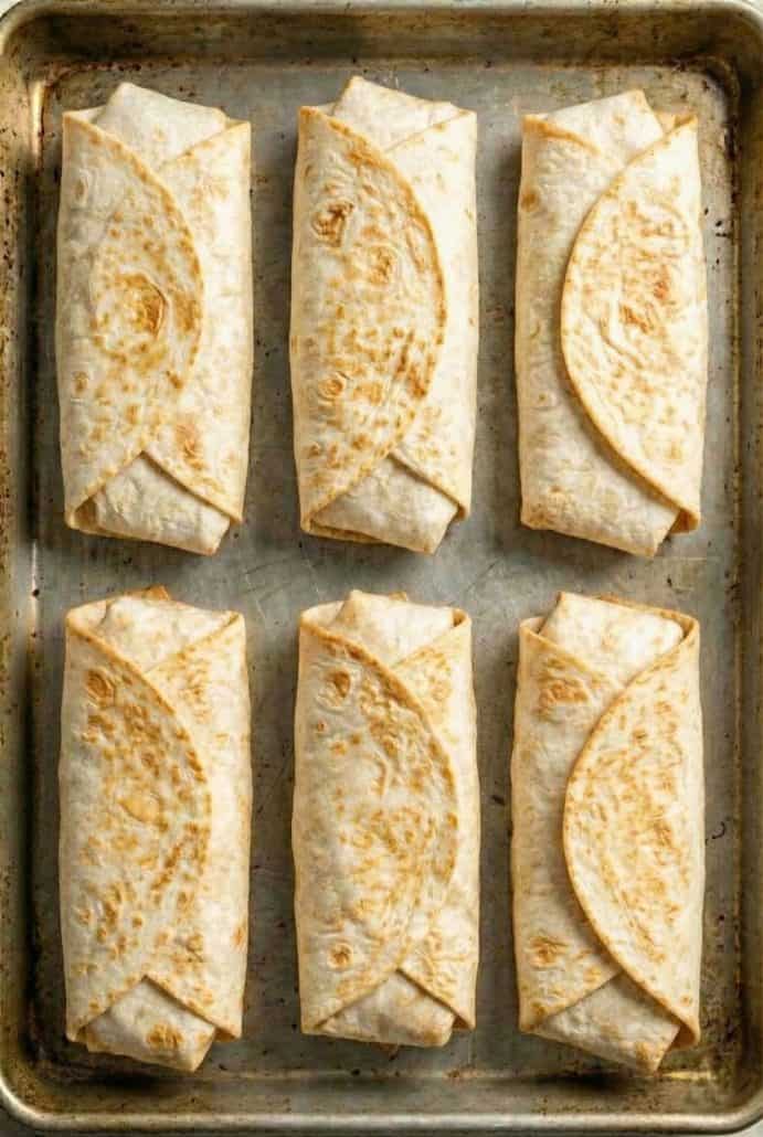 Six rolled sweet potato burritos are arranged neatly on a baking sheet, perfect for meal prep.