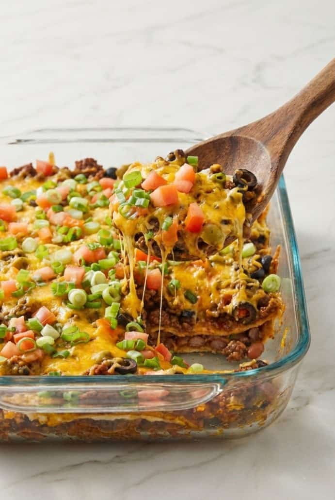Serving a cheesy scoop of taco casserole with ground beef, beans, and toppings from a glass baking dish.