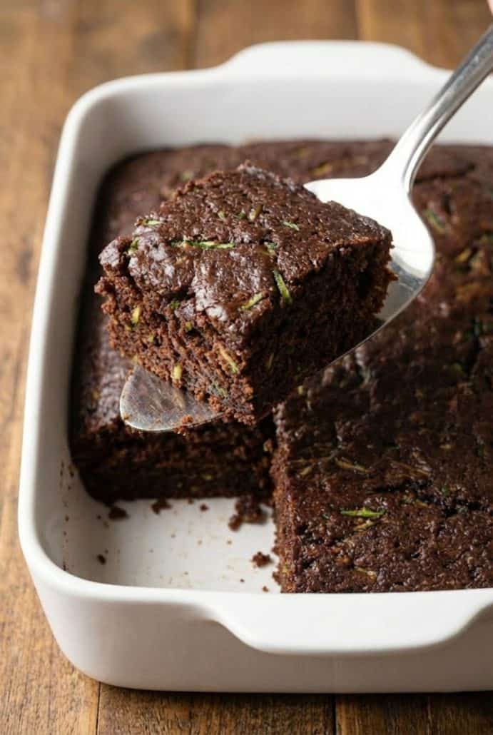 A slice of moist chocolate zucchini cake being lifted from a baking dish, showing the rich, fudgy texture inside.