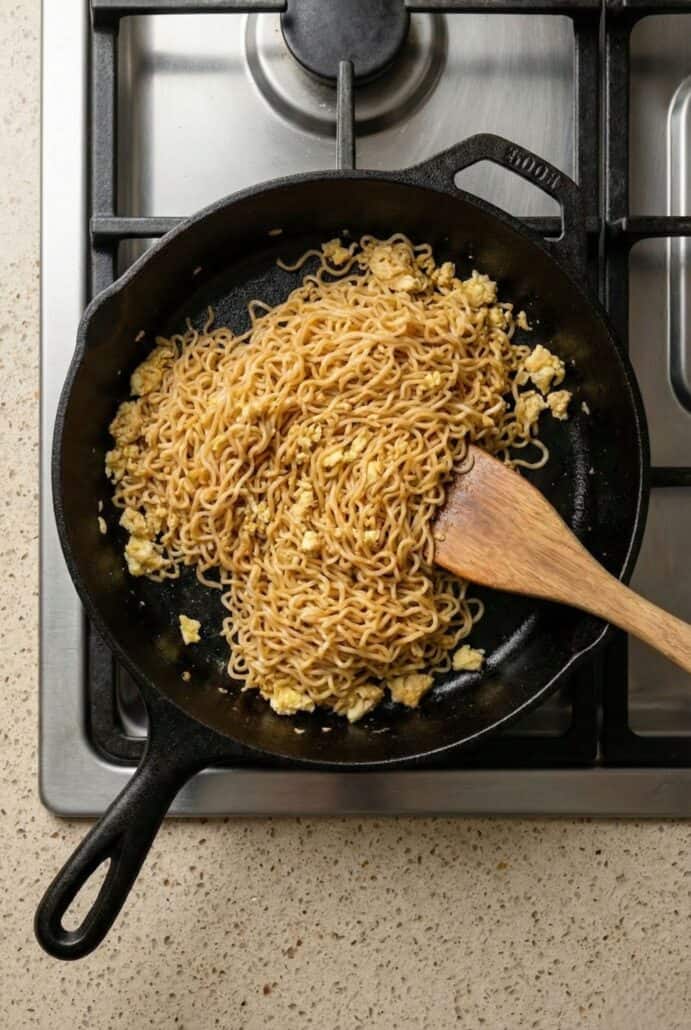 TikTok Ramen cooking in a cast iron skillet with scrambled egg, stirred with a wooden spatula on the stovetop – step-by-step process of making viral ramen noodles.