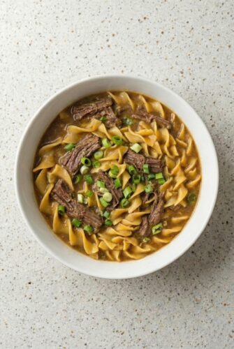 Authentic Amish Beef and Noodles That Tastes Like Home Close-up bowl of Amish beef and egg noodles in rich savory broth, topped with sliced green onions.