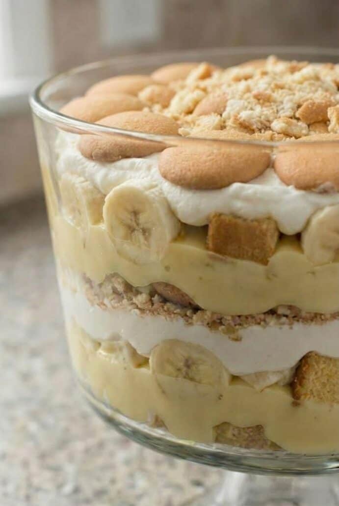 Close-up of banana trifle in a glass bowl showing layers of banana pudding, sliced bananas, pound cake, and vanilla wafers for this easy no-bake dessert recipe.