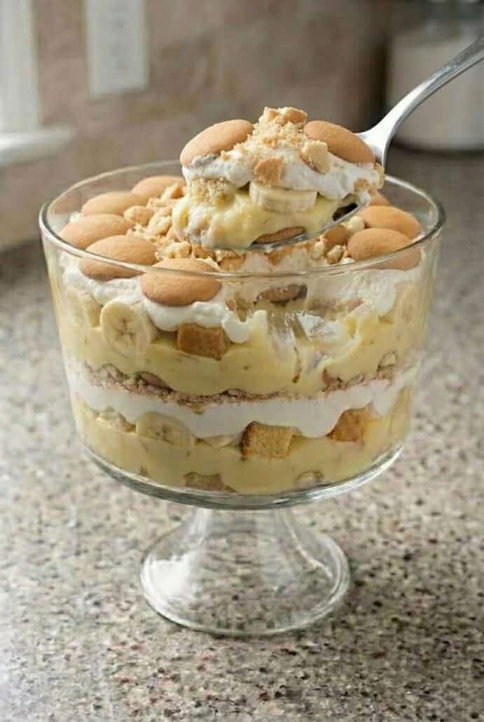 Spoon scooping a serving of layered banana trifle with whipped cream, pudding, pound cake, and vanilla wafers.