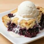 Slice of homemade blackberry cobbler with a golden cake-like topping and a scoop of vanilla ice cream, showing the juicy berry filling.