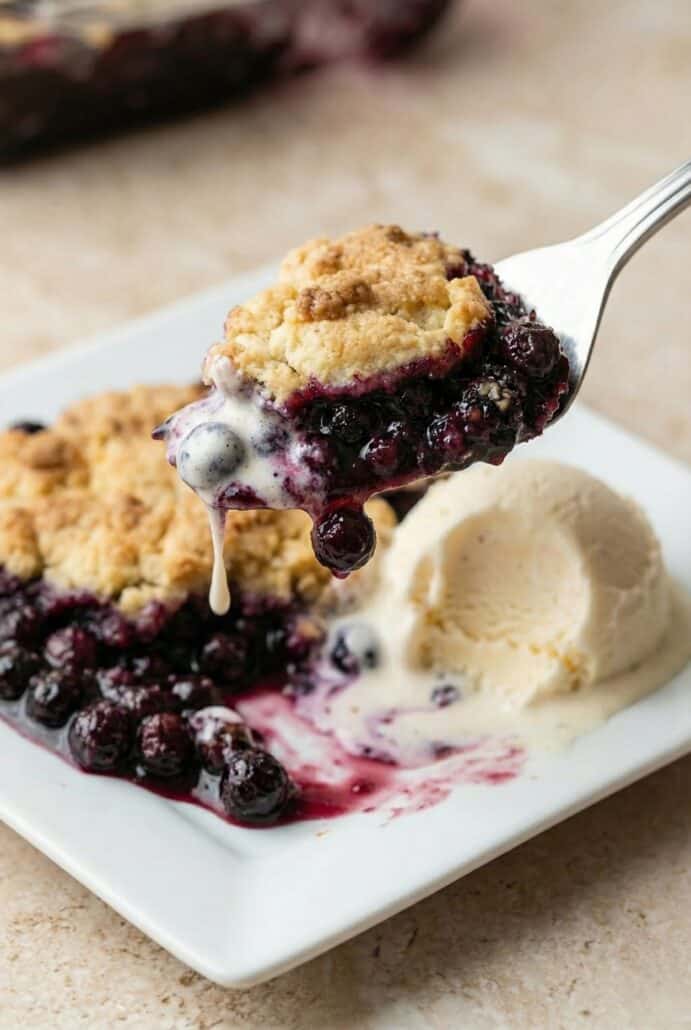 Close-up of blueberry cobbler on a white plate with melting vanilla ice cream, showcasing the juicy berry filling and golden baked topping from this easy homemade cobbler recipe.