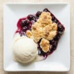 Blueberry cobbler served warm with a scoop of vanilla ice cream, featuring a golden cake-like topping and rich berry filling.