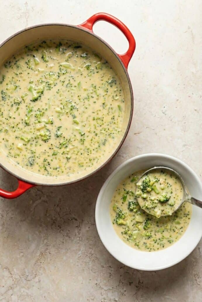Overhead view of creamy broccoli cheese soup in a red pot with a bowl served alongside, showcasing the rich, cheesy texture.