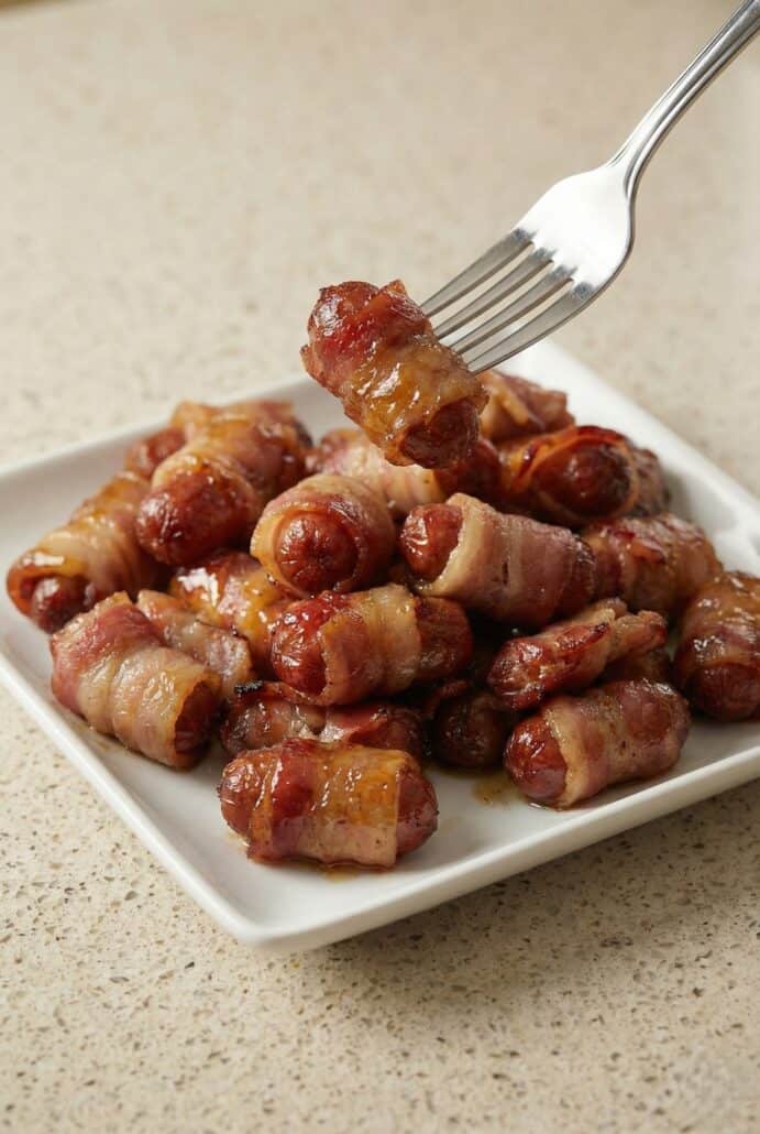 Fork lifting a bacon-wrapped little smokie coated in caramelized brown sugar glaze from a serving plate, highlighting the sticky sweet and savory texture of this easy party appetizer recipe