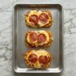 Overhead view of three crispy hashbrown pizzas topped with melted mozzarella and pepperoni on a baking sheet.