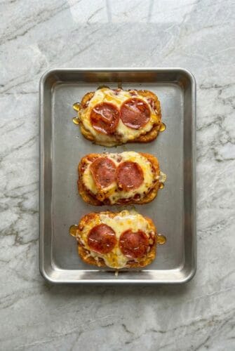 Overhead view of three crispy hashbrown pizzas topped with melted mozzarella and pepperoni on a baking sheet.