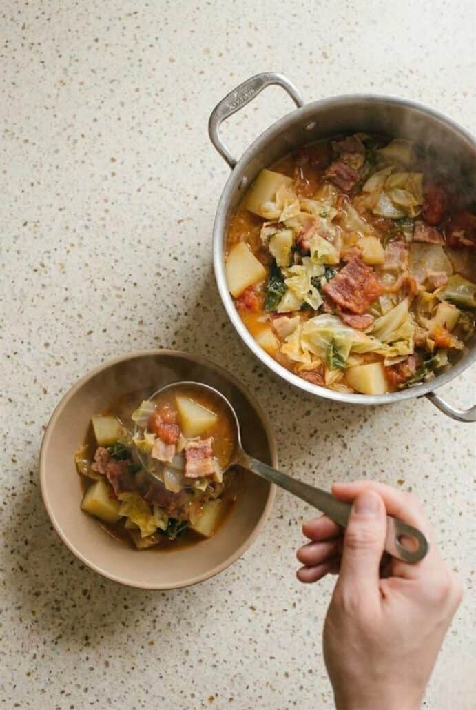 Hand ladling chunky Irish bacon and cabbage soup from a large pot into a bowl, highlighting the tender potatoes, tomatoes, and savory bacon.