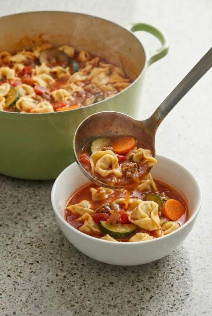 Ladle serving Italian sausage tortellini soup into a bowl, showing hearty vegetables and rich tomato broth.