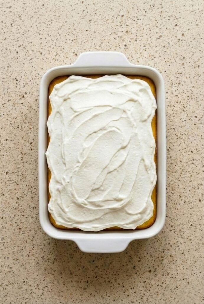 Lemon poke cake in a baking dish topped with smooth white frosting, ready to slice and serve.