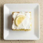 Slice of lemon poke cake topped with white frosting and a fresh lemon slice.