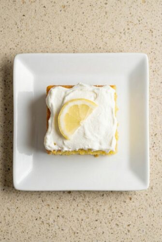 Sweet and Tangy Lemon Poke Cake Recipe Slice of lemon poke cake topped with white frosting and a fresh lemon slice.