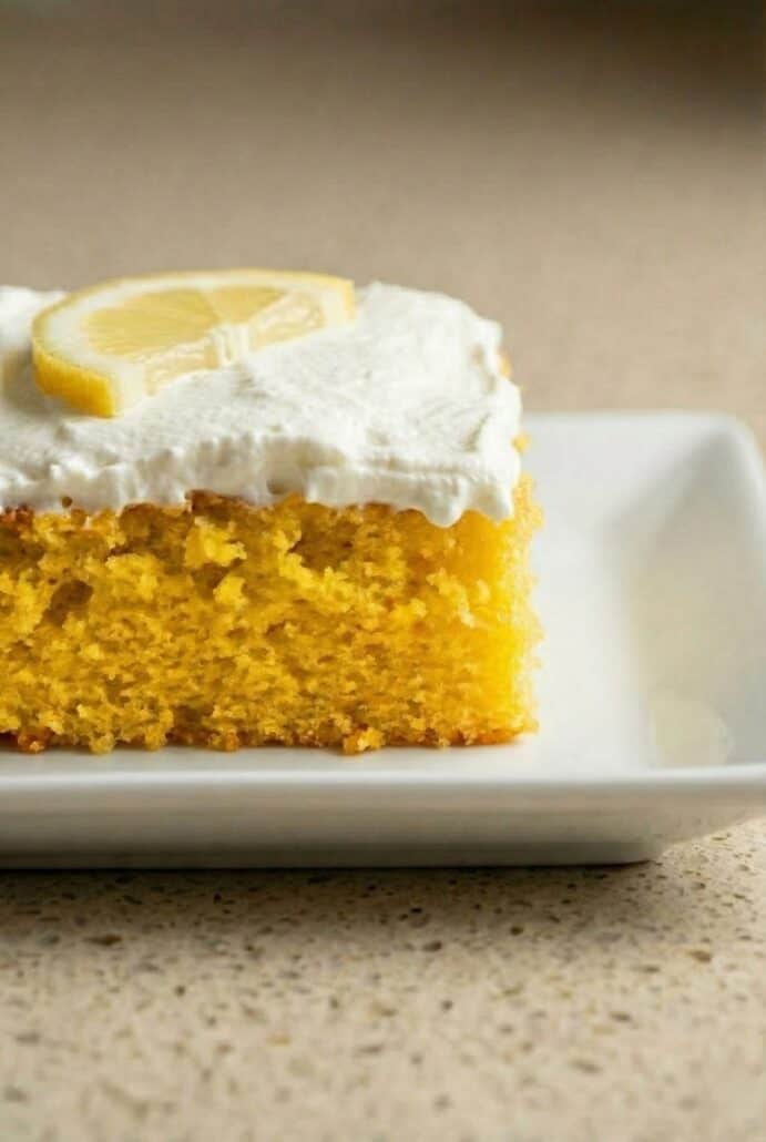 Close-up of a moist slice of lemon poke cake with creamy white frosting and a fresh lemon wedge on top.