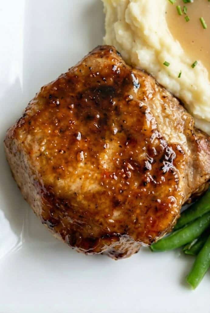 Close-up of glazed baked pork chop plated with mashed potatoes and green beans, highlighting the caramelized sweet and savory marinade.