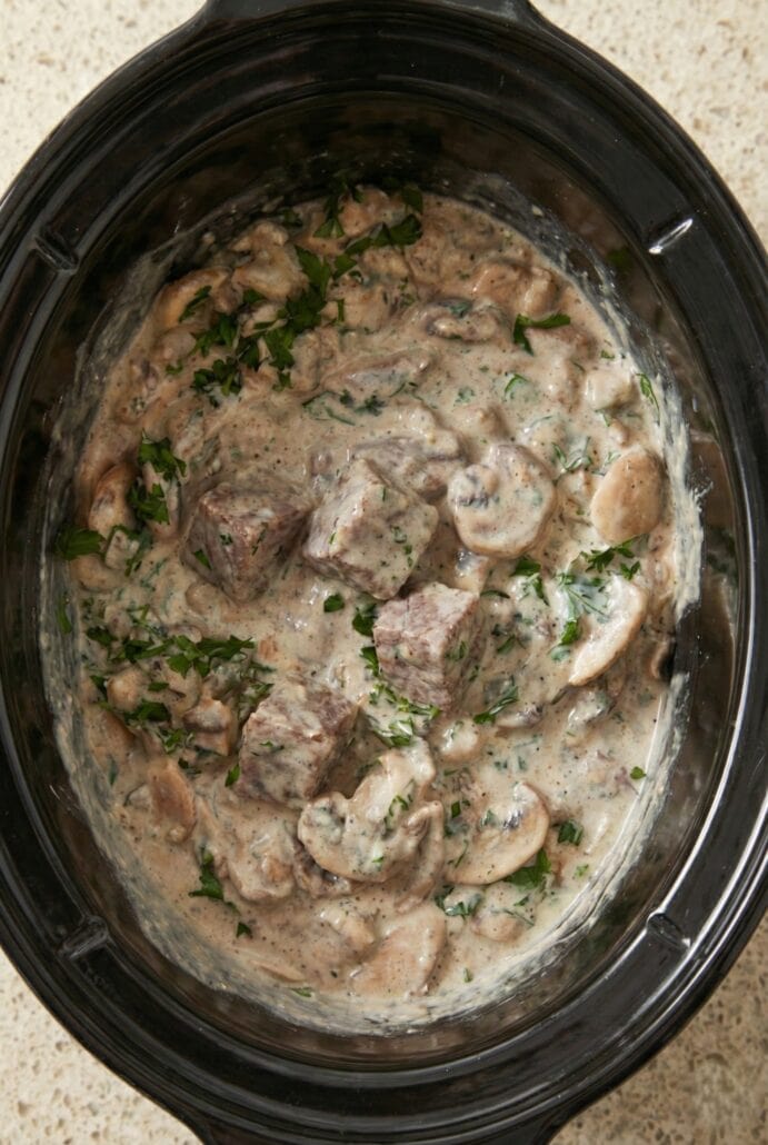 Creamy beef stroganoff cooking in the slow cooker, with tender beef pieces and mushrooms in a rich, creamy mushroom sauce garnished with fresh parsley.