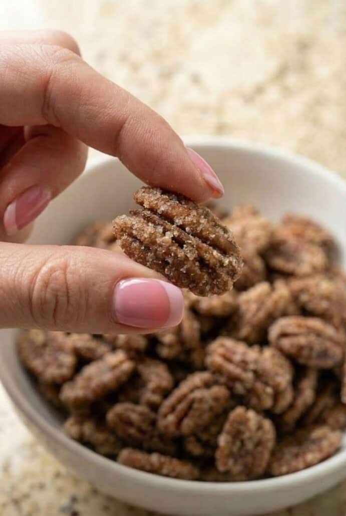 Hand holding a crunchy cinnamon glazed pecan over a bowl, showing the crisp sugary coating from this easy Texas candied pecans recipe.