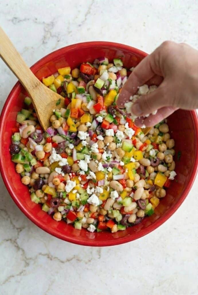 Sprinkling feta cheese over TikTok Dense Bean Salad in a red bowl, just before chilling for enhanced flavor.