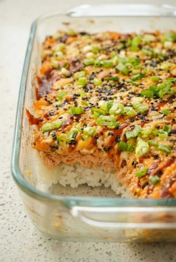 Side view of a TikTok Salmon Sushi Bake in a glass dish with a slice removed, showing layers of sushi rice, creamy salmon topping, furikake, and green onions.