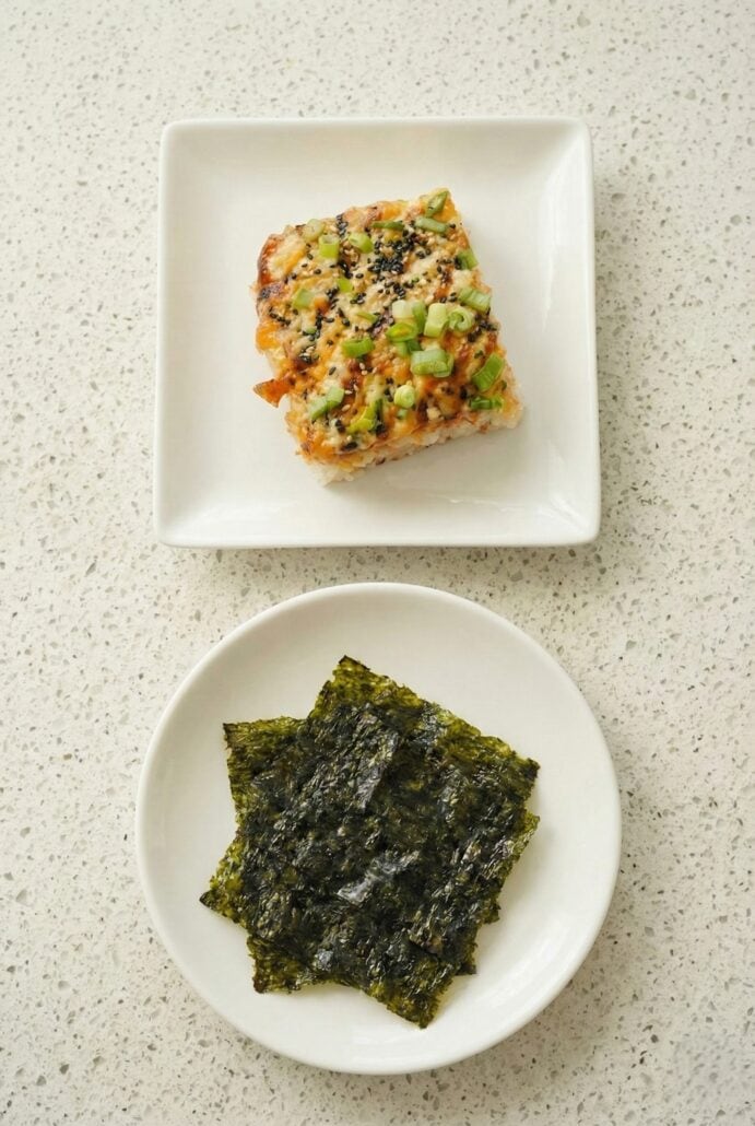 Top-down view of a serving of TikTok Salmon Sushi Bake on a square plate next to a round plate of roasted seaweed snacks, ready to be wrapped and eaten.