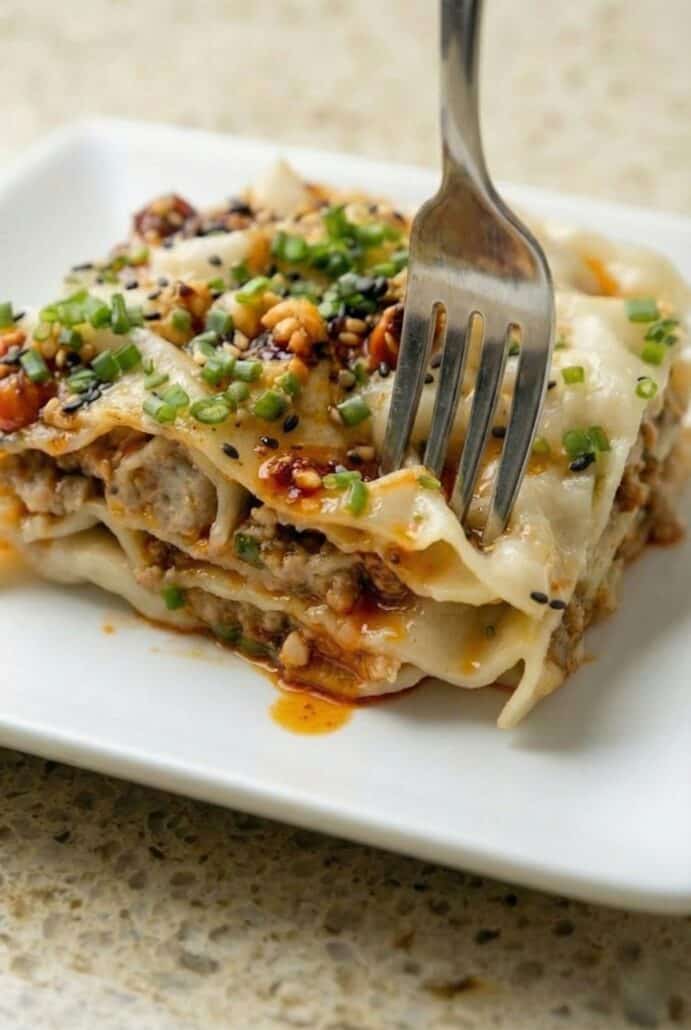 Close-up of a fork lifting a layered piece of viral dumpling lasagna, showing tender dumpling wrappers, savory chicken filling, and a drizzle of sesame sauce with chili oil and chives.
