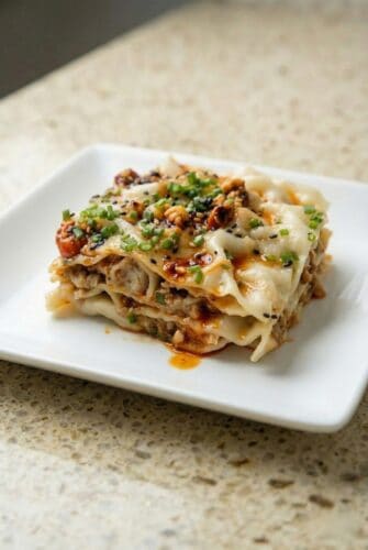 Slice of viral dumpling lasagna layered with dumpling wrappers and savory chicken filling, topped with sesame sauce and chili oil.